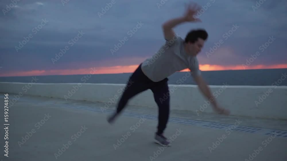 Young Caucasian male gymnast in casual outfit perfoming serie of acrobatic exercises in slowmotion. Beautiful sunset over sea is on background Young Caucasian male gymnast in casual outfit perfoming serie of acrobatic exercises in slowmotion. Beautiful sunset over sea is on background
