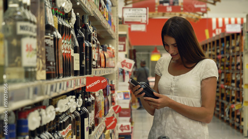 A beautiful young girl (woman) at the grocery store, in a white tank top, supermarket, chooses alcoholic drinks products, with a cart. Concept: buy products, large purchases, market, store, products. © Kitreel