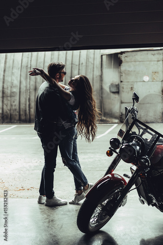 Stylish young couple in love hugging while standing in garage with motorbike