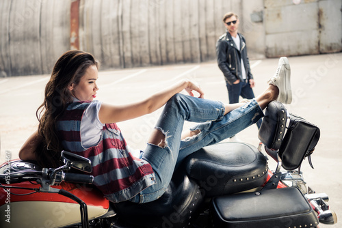 Beautiful young woman in denim vest sitting on motorcycle and stylish man in sunglasses standing behind
