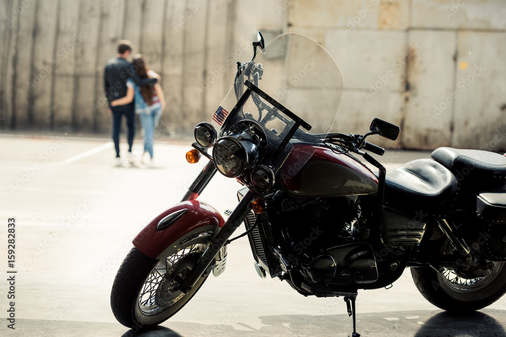 Close-up view of motorcycle standing on pavement and young couple walking behind Close-up view of motorcycle standing on pavement and young couple walking behind