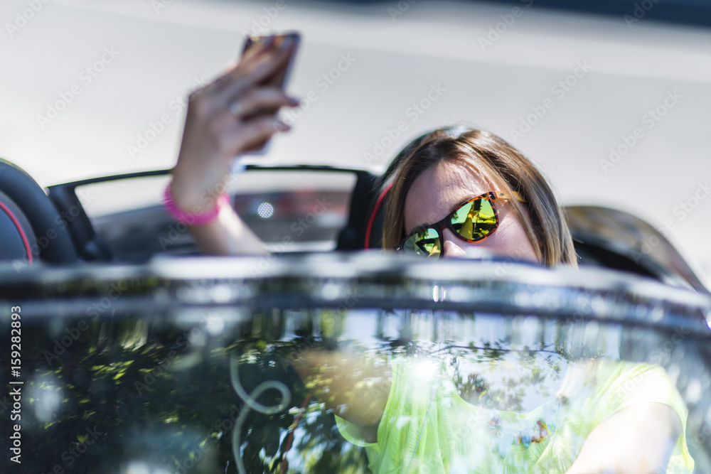 Φωτογραφία Young woman driving a cabriolet car while distracted making a selfie with her smartphone and being dangerous for the traffic around