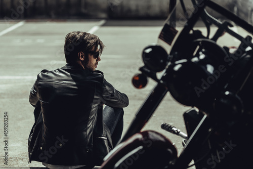 Back view of stylish young man in leather jacket sitting near motorbike and looking away photo