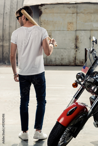 Back view of stylish young man with baseball bat on shoulder standing near motorbike and looking away