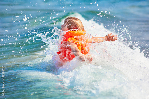 Billede på lærred Little boy in orange life vest swimming in wave sea (selective DOF)
