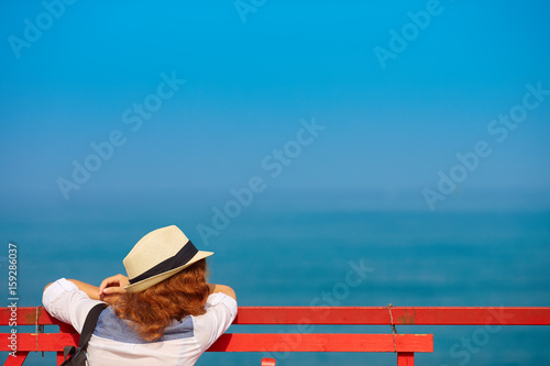 Young redhead woman in hat on pier looks out into ocean, with copy space