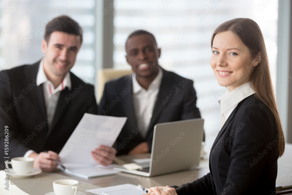 Attractive young businesswoman smiling for camera sitting at desk, young executive manager holding business meeting for black and white subordinates clients at background, female team lead portrait Attractive young businesswoman smiling for camera sitting at desk, young executive manager holding business meeting for black and white subordinates clients at background, female team lead portrait