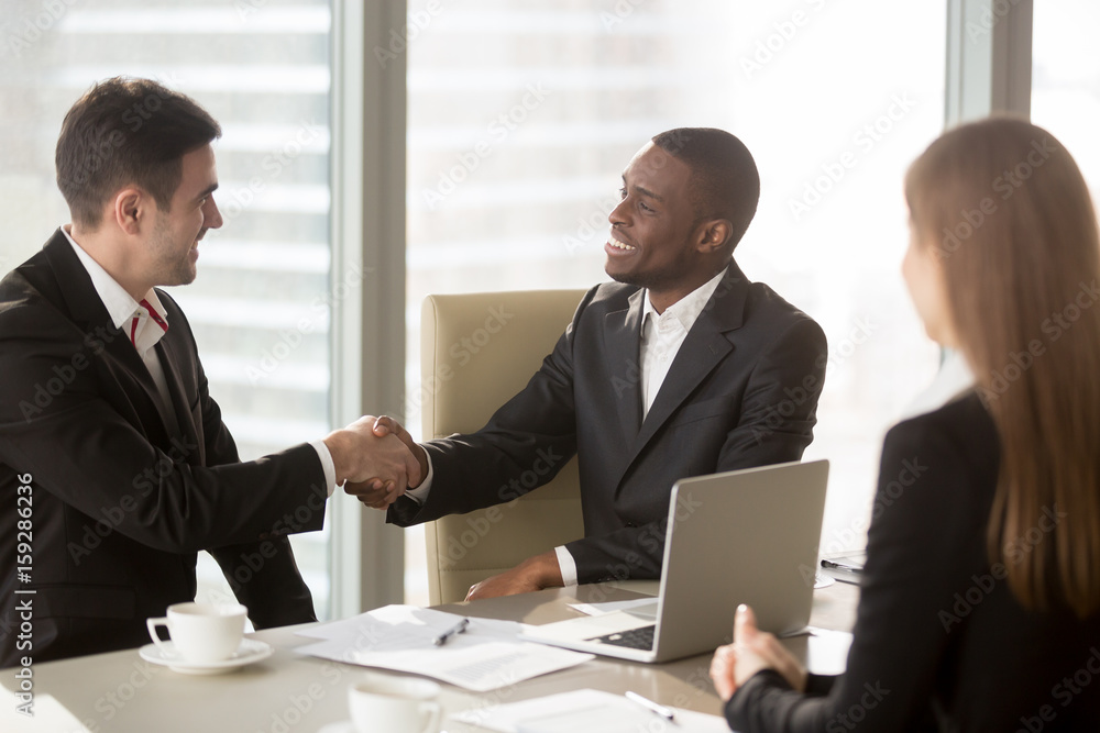 Two cheerful businessmen handshaking at formal meeting, starting finishing negotiations, african entrepreneur wearing black suit white shirt and caucasian partner shake hands, making profitable deal Two cheerful businessmen handshaking at formal meeting, starting finishing negotiations, african entrepreneur wearing black suit white shirt and caucasian partner shake hands, making profitable deal
