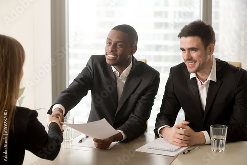 Two cheerful black and white recruiters welcoming female applicant on job interview, african and caucasian hr managers greeting candidate for vacant position, handshaking and good first impression