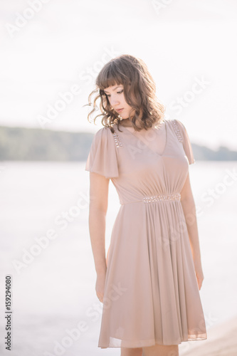 very beautiful, sexy, gentle, cute girl in beige dress stands on the banks of the river, outdoors in summer smiling, looking down, profile, close-up