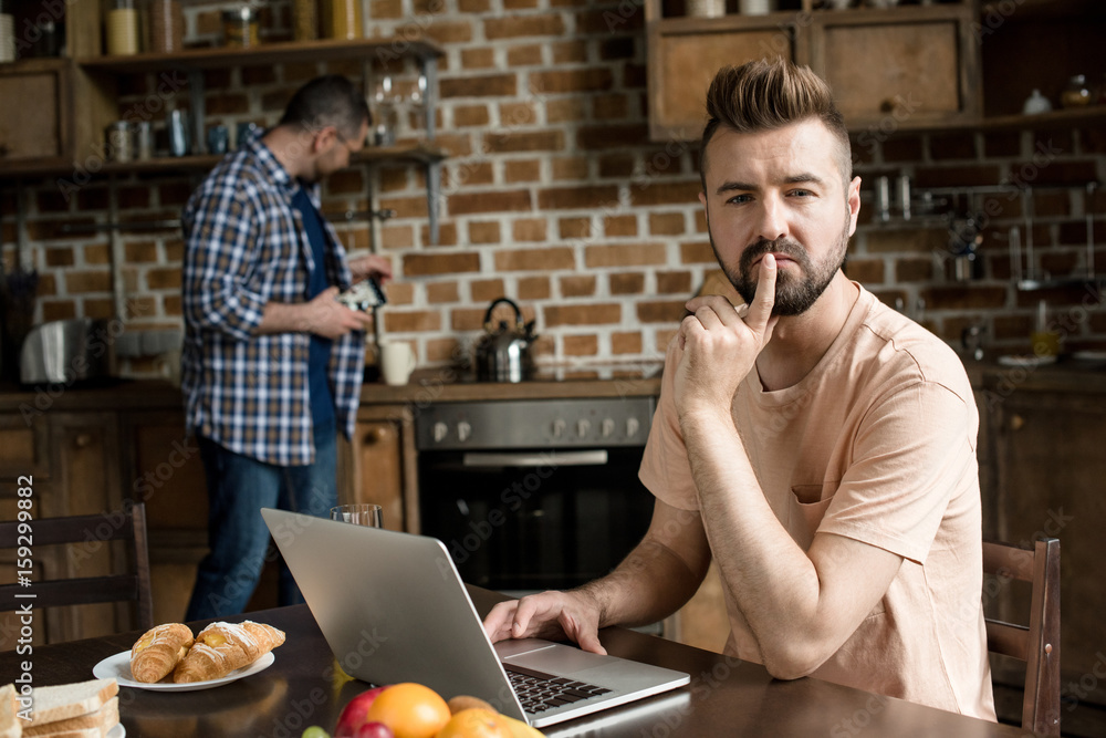 Thoughtful bearded man sitting at table and using laptop while partner pouring coffee behind