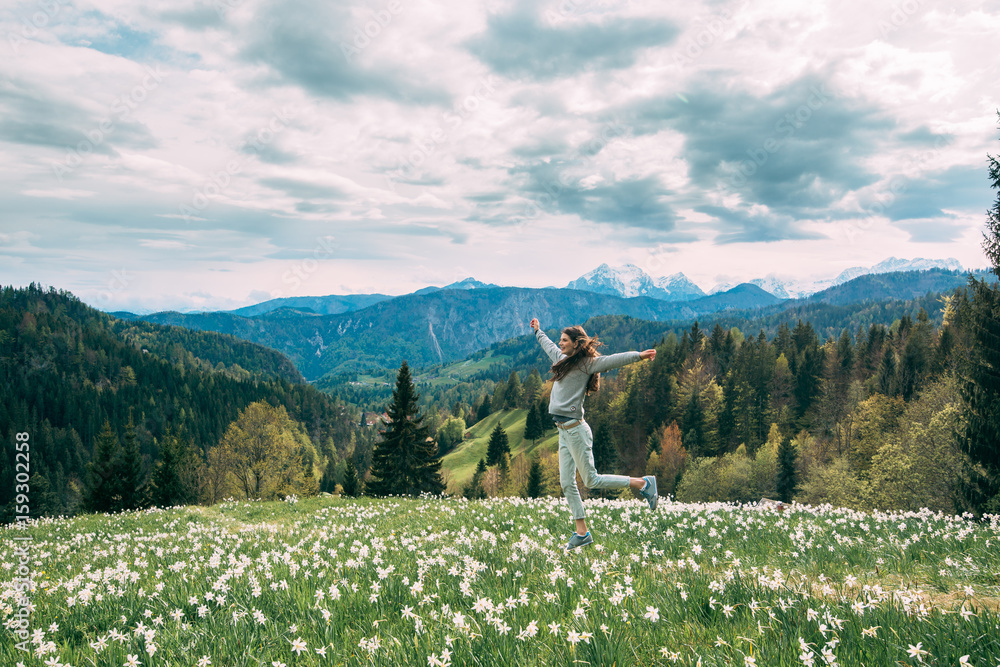 Fototapeta premium Young woman jumping in daffodil field, blooming narcissus with mountains on background