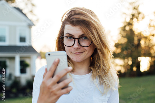 Pretty attractive female with blonde hair and round nerdy glasses makes a selfie photo portrait with smartphone, looks at reflection in screen, judging her appearance photo