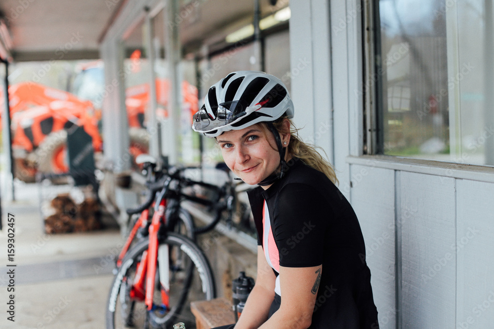 Tired but happy, exhausted after long training ride female rider smiles into camera in her helmet and lycra cycling kit, with professional road bikes in distance Tired but happy, exhausted after long training ride female rider smiles into camera in her helmet and lycra cycling kit, with professional road bikes in distance