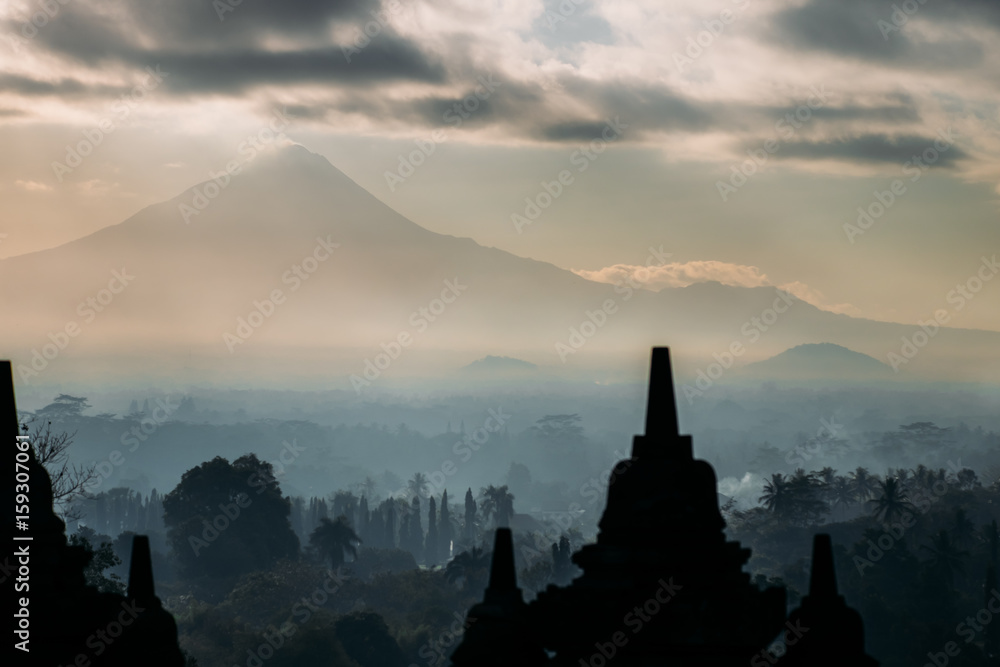 Borobudur in the morning during sunrise. View of foggy forest during the sunrise. The mist covers all trees and forest below the temple. Big volcano mount Marapi is in under the morning sky and fog. 
