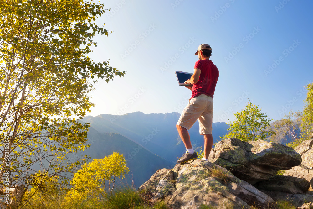 Young caucasian man sitting outdoor on a rock working on a laptop pc in mountain area.