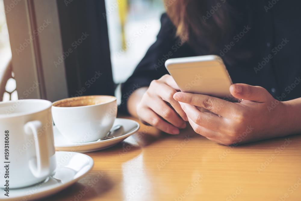 A woman with smiley face holding and using smart phone with coffee cup on wooden table in the cafe A woman with smiley face holding and using smart phone with coffee cup on wooden table in the cafe