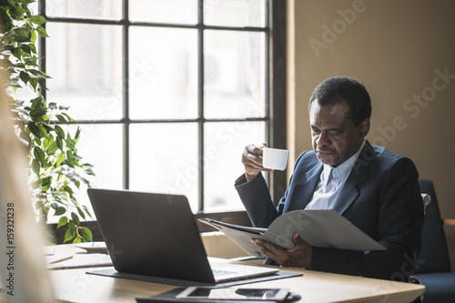 Middle-aged African businessman sitting at office and reading and holding cup of coffee.