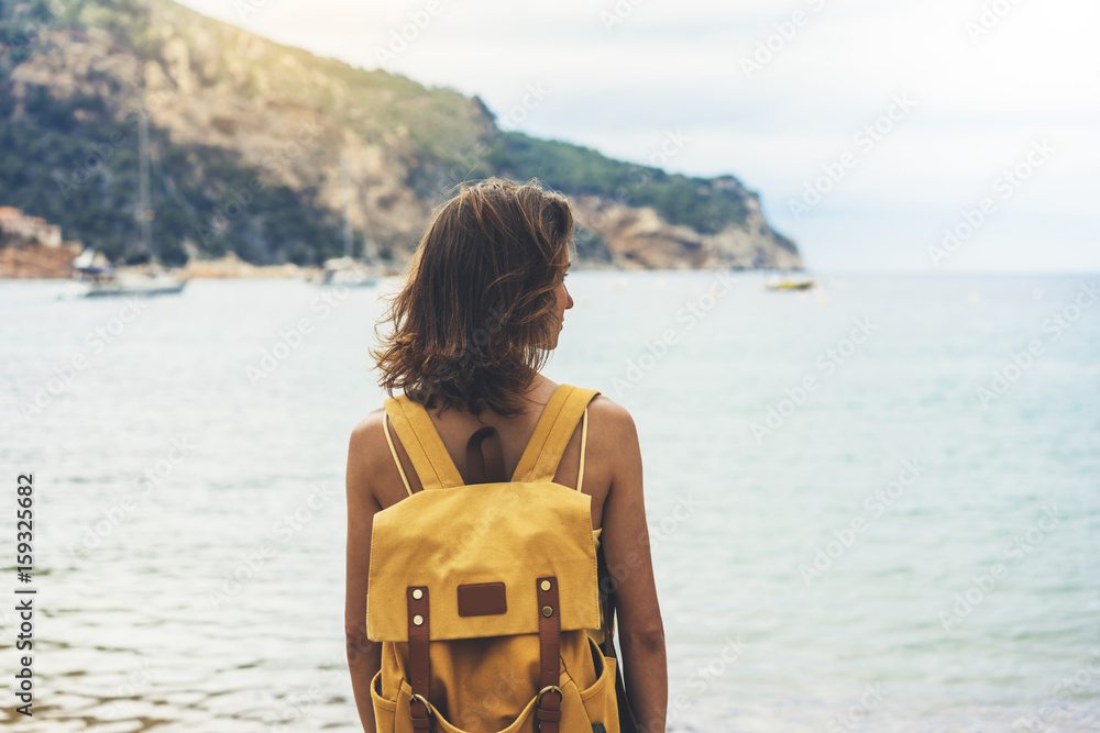 Fototapeta premium Back view hipster girl with backpack in sand coastline on nature landscape, mock up. Traveler on background beach seascape and horizon mountain. Tourist look on blue sun ocean, summer relax lifestyle