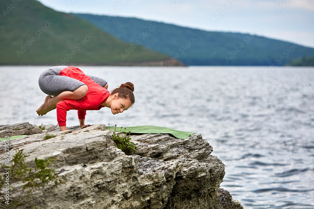 Young woman doing yoga pose arm balance on the stone near big river.