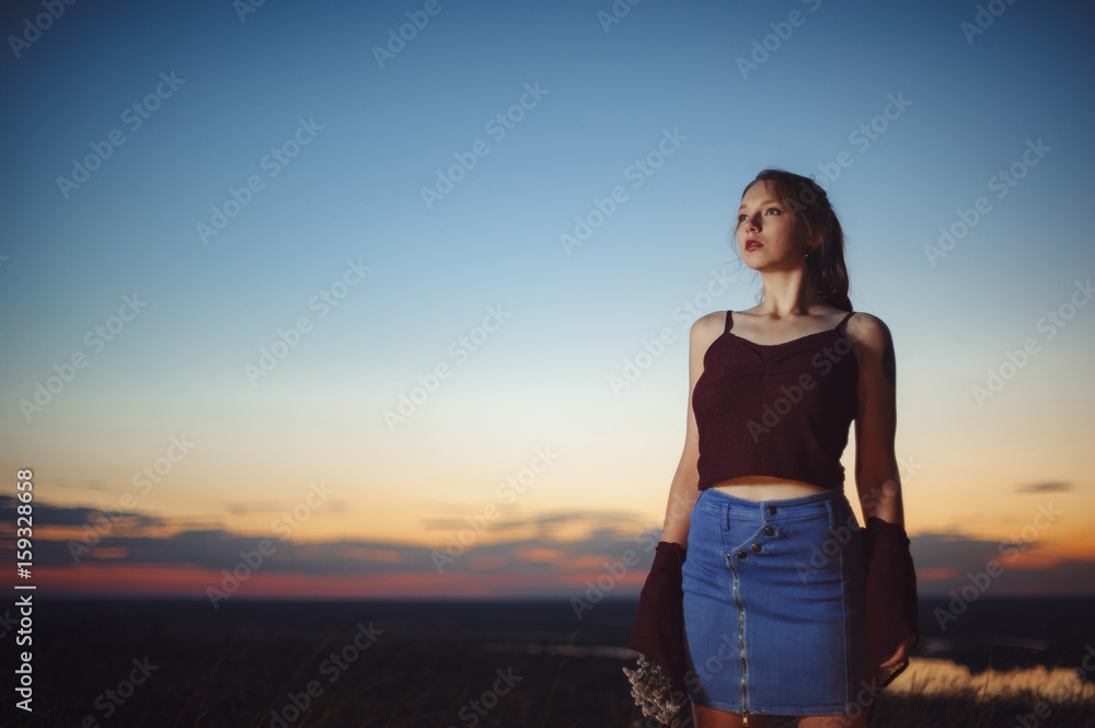 Red-haired girl is standing on the brink of a precipice against the backdrop of a beautiful sunset sky. Grunge style of clothes. Flowers in hand. Red-haired girl is standing on the brink of a precipice against the backdrop of a beautiful sunset sky. Grunge style of clothes. Flowers in hand.