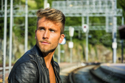 Attractive blond young man standing on railroad tracks, wearing only black leather jacket, looking away