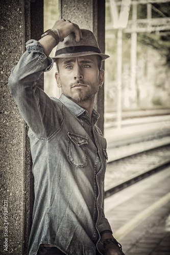 Portrait of blond haired handsome young man with fedora hat. An old train station is behind him in Italian town © starsstudio