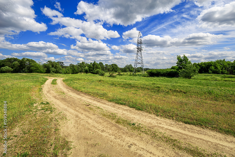 road and green field with trees and blue sky with clouds sunny day, beautiful rural landscape. Electricity tower standard overhead power line transmission tower road and green field with trees and blue sky with clouds sunny day, beautiful rural landscape. Electricity tower standard overhead power line transmission tower