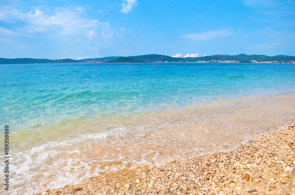 Pebble sandy beach on the background of a hill range with many small houses, a line of buoys and azure transparent blue sea. Blue cloudless sky. Zadar, Croatia Pebble sandy beach on the background of a hill range with many small houses, a line of buoys and azure transparent blue sea. Blue cloudless sky. Zadar, Croatia
