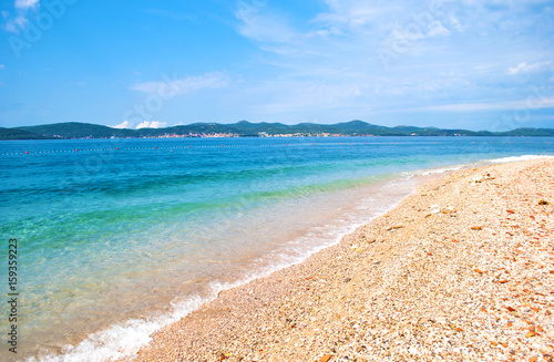 Pebble sandy beach on the background of a hill range with many small houses, a line of buoys and azure transparent blue sea. Blue cloudless sky. Zadar, Croatia
