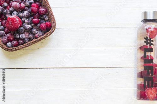 Bottle with refreshing drink, water with strawberry slices, with hashtag #life and basket with frozen berries on white wooden background