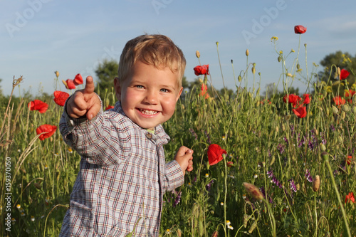 A small blond boy with a toddler in a plaid shirt with a red poppy in his hand. Emotional happy laughing kid runs half a flower in his hand. Farmer boy holding a red poppy in his hand