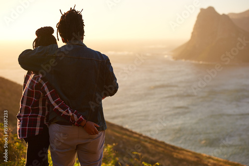 Foto Casually dressed mixed race couple embracing each other while enjoying the awesome view of the ocean and mountains in front of them, taken from behind with the womans hand in the mans back pocket