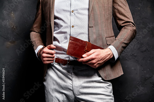 Fashion portrait of young businessman handsome model man dressed in elegant brown suit with accessories on hands posing on gray background in studio. With purse in hands