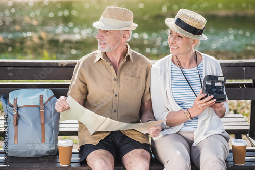 Happy senior couple of travelers sitting together on bench and holding map and instant camera photo
