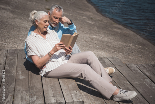 elderly couple reading book while resting on the quay at daytime