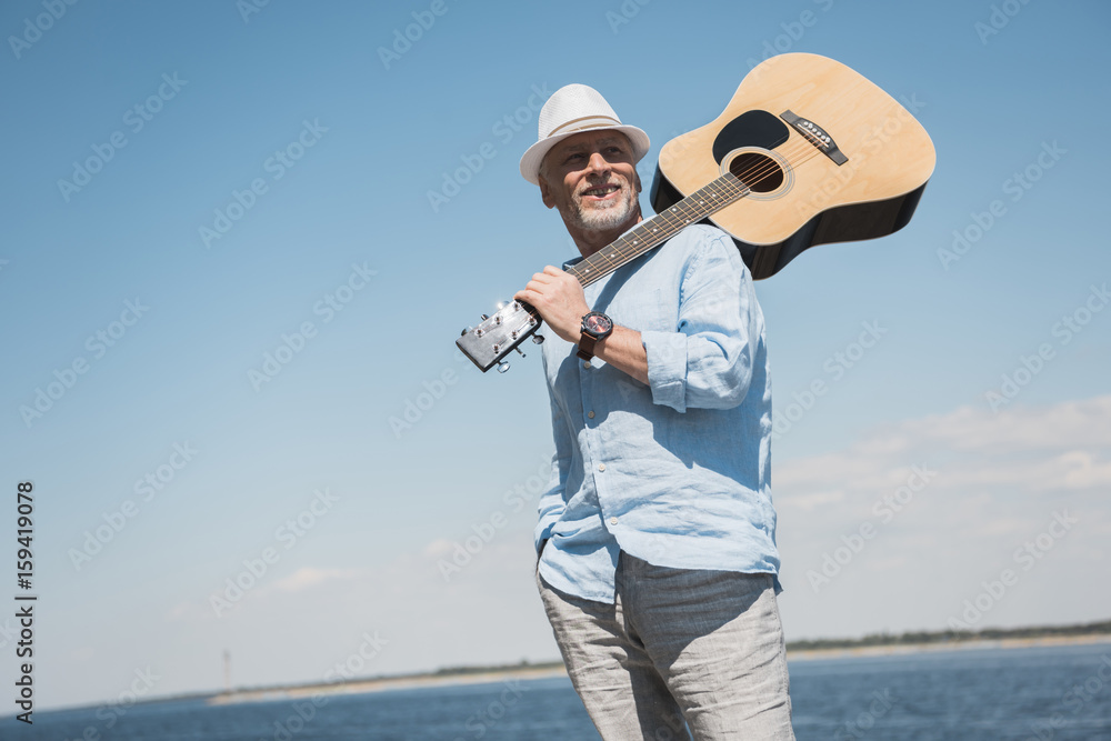 Obraz premium Smiling senior man in hat holding acoustic guitar on shoulder and looking away outdoors
