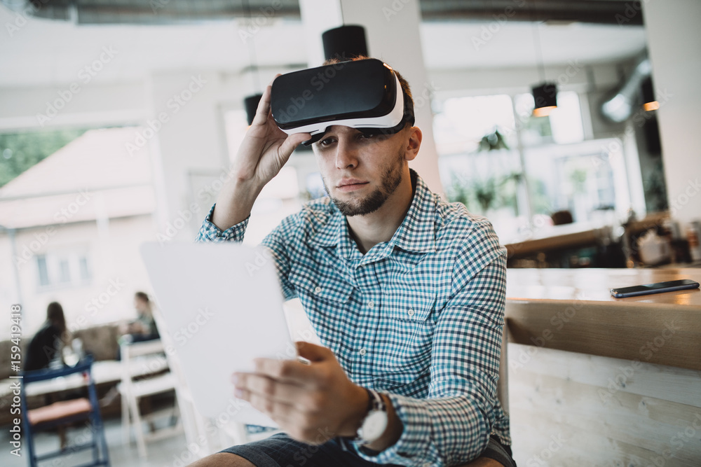 Young handsome man sitting in cyber cafe and using virtual reality headset or glasses. Young handsome man sitting in cyber cafe and using virtual reality headset or glasses.