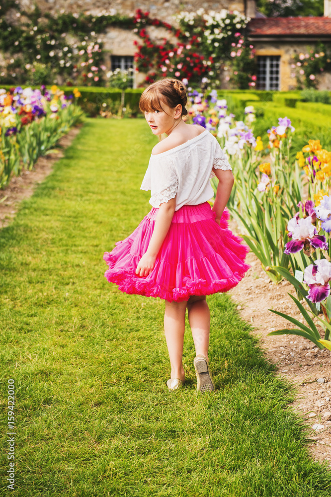 Outdoor portrait of adorable little girl playing in a beautiful flower park on a nice summer day. Fashion kid girl wearing white blouse and bright pink tutu skirt, looking back over shoulder Outdoor portrait of adorable little girl playing in a beautiful flower park on a nice summer day. Fashion kid girl wearing white blouse and bright pink tutu skirt, looking back over shoulder
