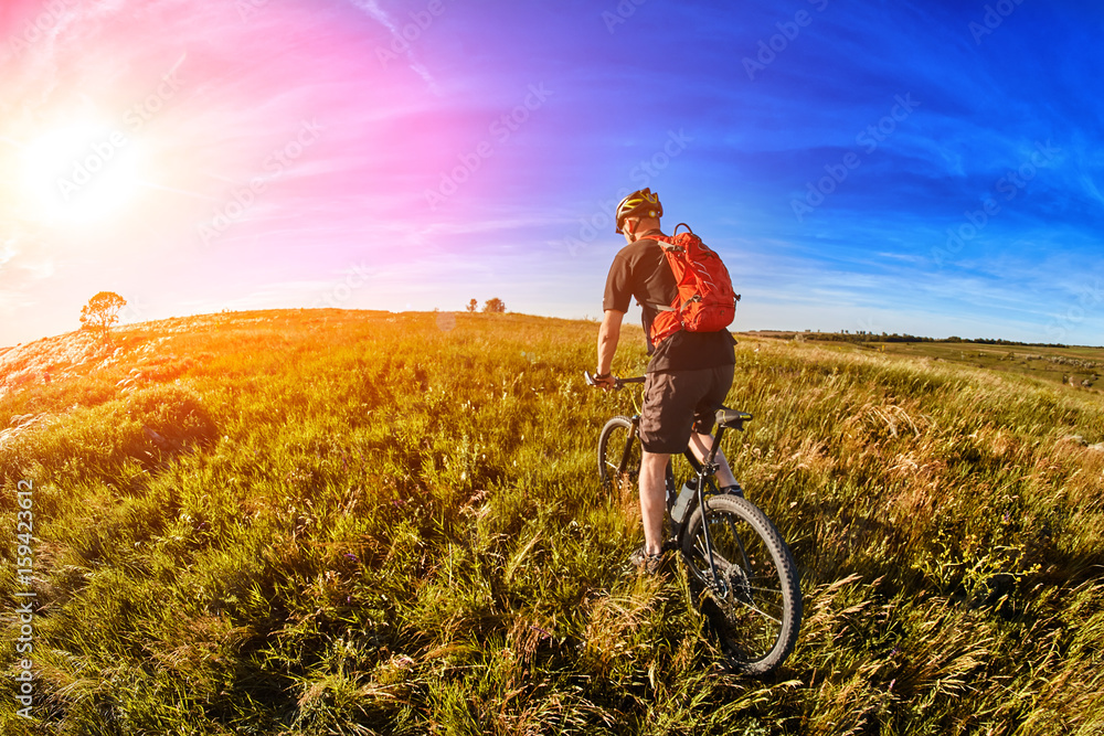 Fototapeta premium Young cyclist riding mountain bicycle through green meadow against beautiful sky.