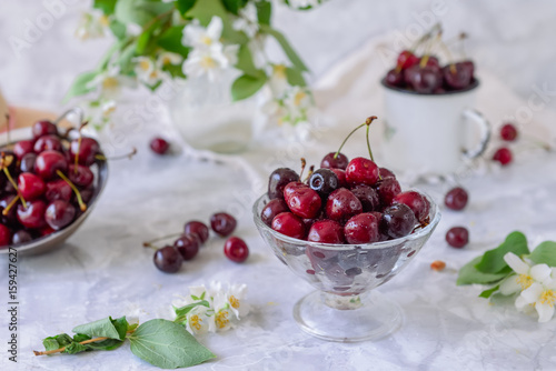 Fresh cherry fruit in glass vase, other dishes with berries and jar with jasmine and wildflowers on the light marble table. Soft selective focus. Summer countryside concept