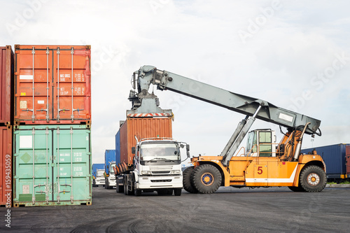 Forklift truck lifting cargo container in shipping yard from trailer truck at dock yard with cargo container stack in background for transportation import, export and logistic industrial concept
