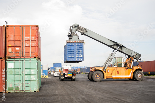 Forklift truck lifting cargo container in shipping yard from trailer truck at dock yard with cargo container stack in background for transportation import, export and logistic industrial concept
