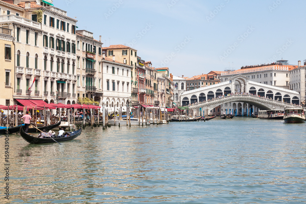 Obraz premium June 2017 Rialto Bridge, Venice, Italy. Gondolas and vaporettos on the Grand Canal with reflections from historic palazzos on the water