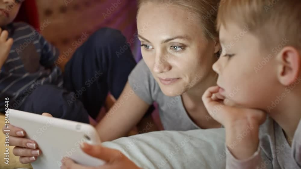 Closeup of mother lying on floor, using digital tablet and telling something to little sons Closeup of mother lying on floor, using digital tablet and telling something to little sons