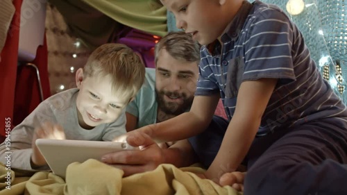 Tracking shot of father and two little sons lying on carpet under play tent decorated with lights and playing game on digital tablet photo