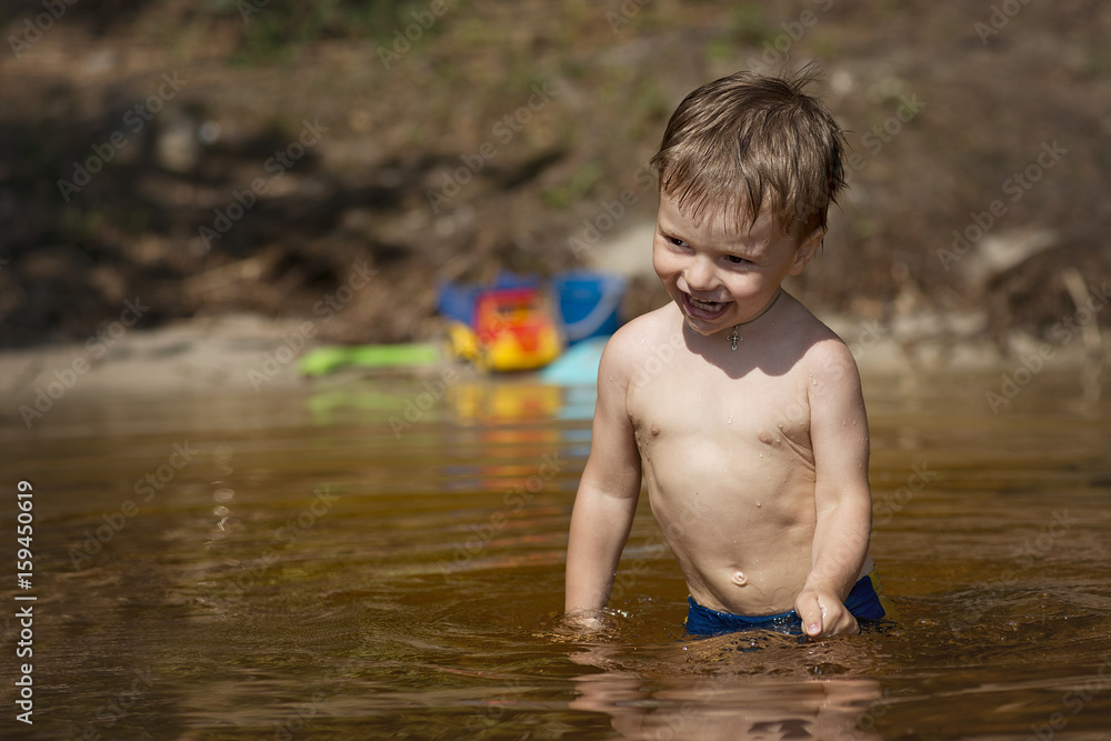 A little boy stands in the water of the lake to the waist and smiling A little boy stands in the water of the lake to the waist and smiling