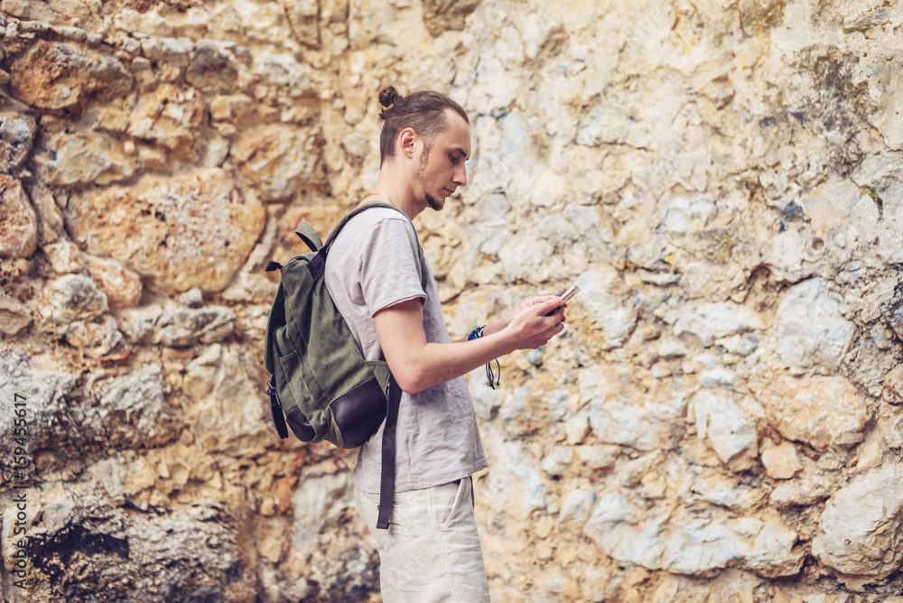 Man traveler with backpack looking at his smartphone on the background of a stone wall