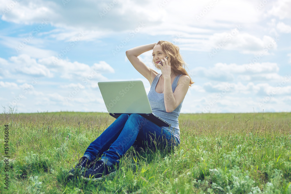 Woman sitting on a green meadow on the background of sky with clouds and working or studying with laptop wireless Woman sitting on a green meadow on the background of sky with clouds and working or studying with laptop wireless