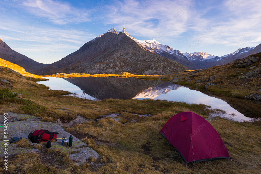 Camping with tent near high altitude lake on the Alps. Reflection of snowcapped mountain range and scenic colorful sky at sunset. Adventure and exploration.